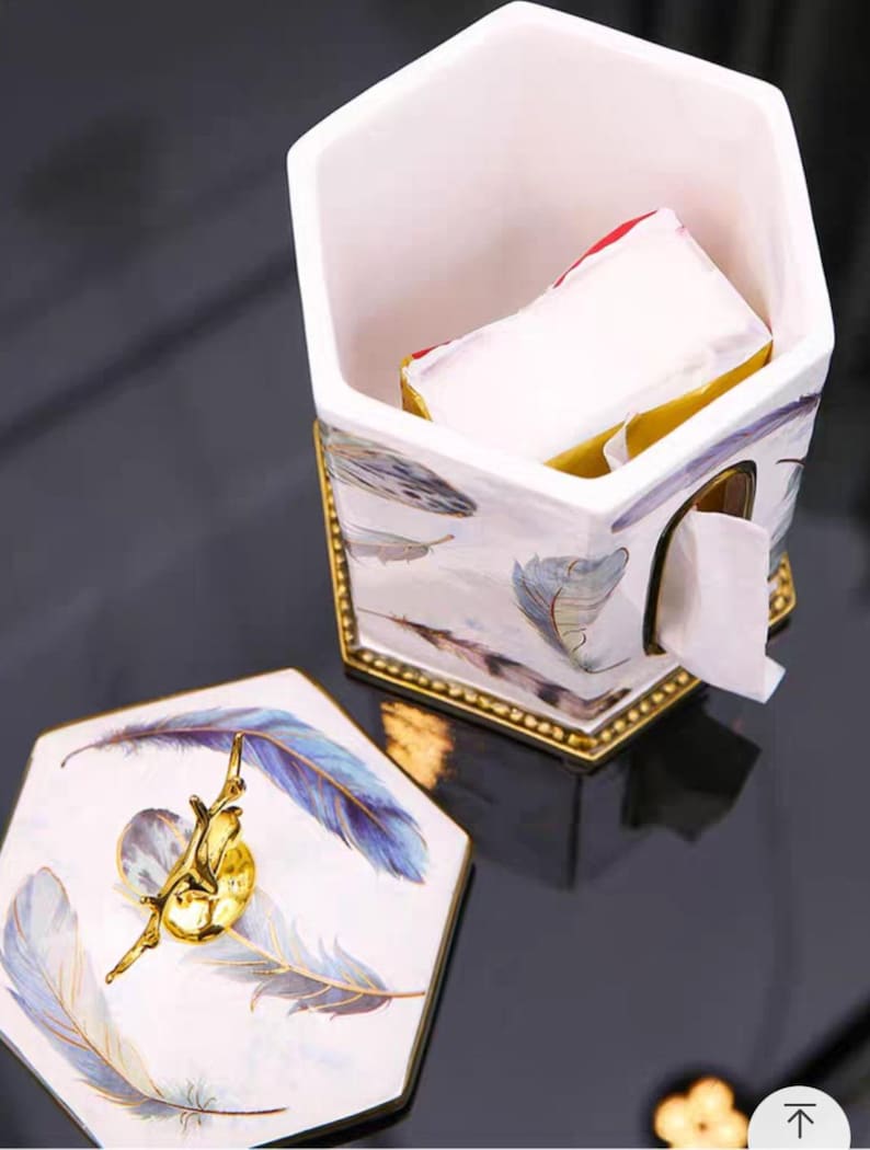 May include: White ceramic tissue box holder with a hexagonal shape and a gold trim. The holder has a lid with a gold knob and a feather design. The holder is filled with white tissues.
