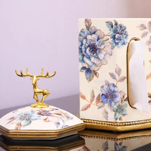 May include: A white ceramic tissue box holder with a floral design in shades of blue and gold. The holder has a gold trim and a deer figurine on top.