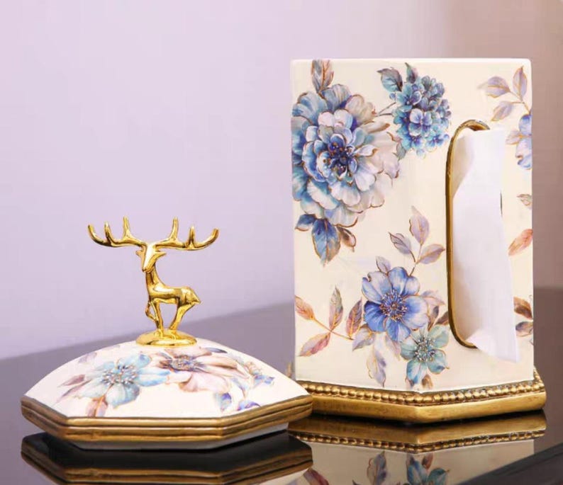 May include: A white ceramic tissue box holder with a floral design in shades of blue and gold. The holder has a gold trim and a deer figurine on top.