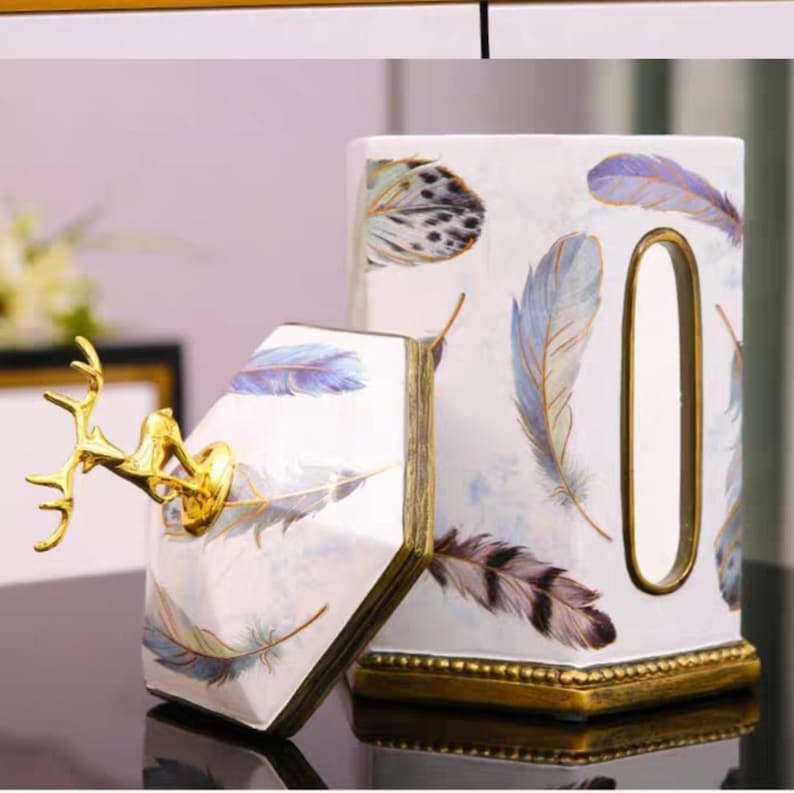 May include: White ceramic tissue box cover with a gold trim and a deer figurine on top. The box is decorated with a pattern of blue and white feathers.