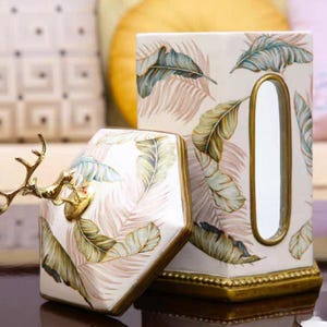 May include: A white ceramic tissue box cover with a gold trim and a tropical leaf design. The cover has a slot for tissues and a decorative deer head on top.