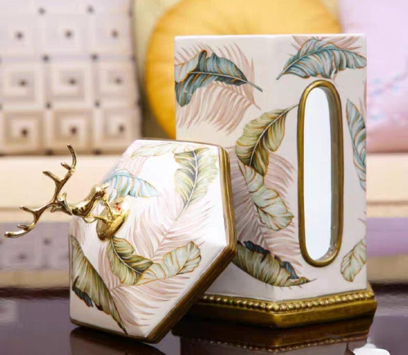 May include: A white ceramic tissue box cover with a gold trim and a tropical leaf design. The cover has a slot for tissues and a decorative deer head on top.
