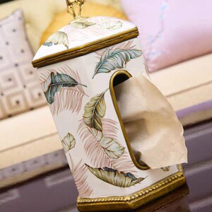 May include: A white ceramic tissue box cover with a gold trim and a tropical leaf design. The cover has a slot for pulling out tissues.