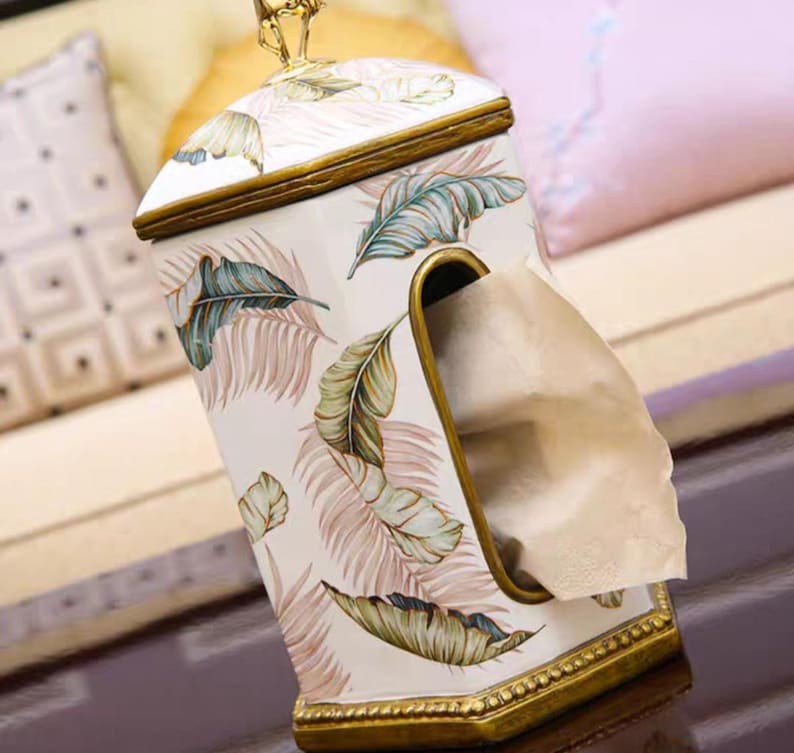 May include: A white ceramic tissue box cover with a gold trim and a tropical leaf design. The cover has a slot for pulling out tissues.