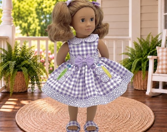 May include: A doll with brown hair styled in two pigtails with purple bows, wearing a purple and white gingham dress featuring a Peter Pan collar, a central purple bow at the waist, two butterfly appliques on the skirt, and matching purple sandals.