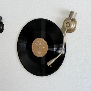 May include: A black vinyl record mounted on a wall with a gold-toned metal tonearm display bracket. The record features a gold centre label with the text Billie Eilish Happier Than Ever. The bracket includes a textured cylindrical knob.