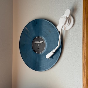 Puede incluir: Un disco de vinilo azul montado en una pared con un brazo y una base de tocadiscos blancos. El disco tiene el texto "HIT ME HARD AND SOFT". La configuración está contra una pared gris claro.