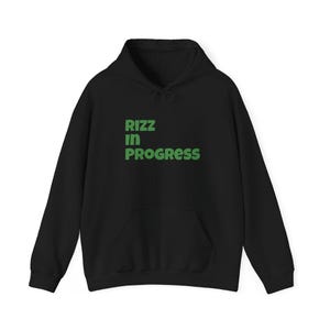 May include: Black hoodie with a front pocket and drawstring. The text "RIZZ IN PROGRESS" is printed in green block letters across the chest. The sweatshirt is made of a soft material.