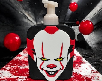 Peut inclure: Distributeur de savon noir avec un visage de clown blanc, des accents rouges et des yeux jaunes. Le distributeur a une pompe blanche et est entouré de ballons rouges. Le design est inspiré d'un film d'horreur.