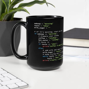 May include: Black coffee mug with white text resembling code. The text includes phrases like "wakeup = 'morning'" and "coffee is good, day". The mug has a large handle and is on a white surface.