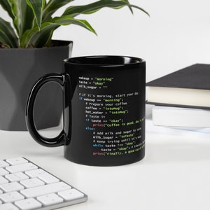 May include: Black coffee mug with a Python code snippet printed on the side. The code includes phrases like "morning", "okay", and "intoMug". The mug is on a white surface with a keyboard and a plant in the background.
