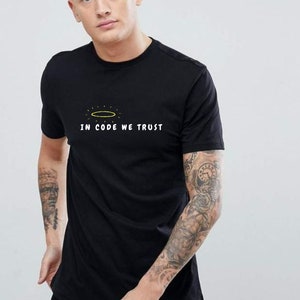 Op de afbeelding: Zwart T-shirt met een witte halo en de tekst "In Code We Trust" gedrukt op de voorkant.