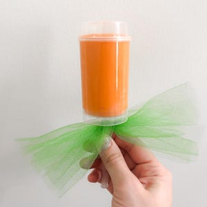 May include: An orange-colored push pop container with a clear lid, held by a hand. The container is attached to a bright green tulle bow. The background is a plain white wall.