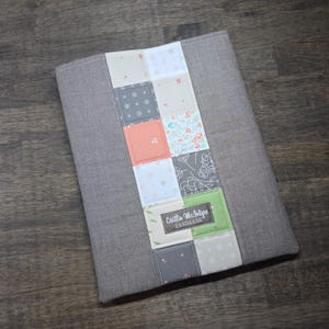 May include: A gray fabric journal cover with a patchwork design down the center. The patchwork includes squares of various patterns and colors, including floral, polka dots, and solid colors. The cover has the text "Caitlin McIntyre Handmade".