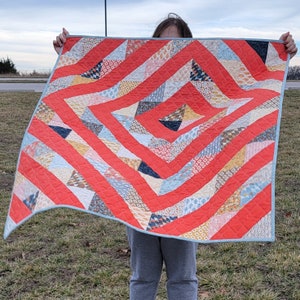 May include: A red, white, and blue quilt with a geometric pattern. The quilt is made of various fabrics and has a light blue border.