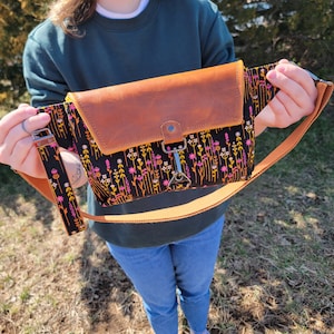 Op de afbeelding: Een bruine leren en zwarte stoffen crossbody tas met een bloemenprint. De tas heeft een klep sluiting en een zilveren gesp. De band is verstelbaar en kan over de schouder of over het lichaam worden gedragen.
