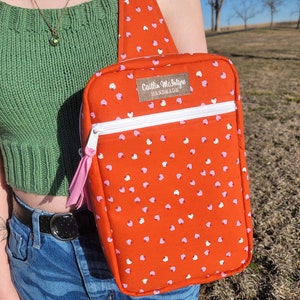 Puede incluir: Una bolsa de bandolera roja con un patrón de corazones blancos y rosas. La bolsa tiene una cremallera blanca y un tirador de cremallera rosa. La bolsa tiene una etiqueta que dice "Caitlin McIntyre Handmade".