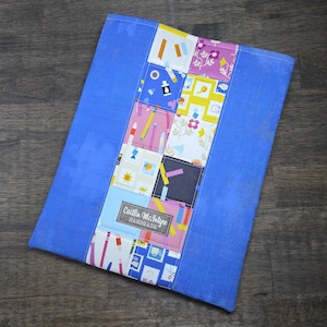 Peut inclure: Un article en tissu bleu fait à la main avec un motif patchwork central. Le patchwork présente divers motifs colorés, notamment des crayons, des timbres et des motifs floraux. Une étiquette indique "Caitlin McIntyre Handmade."