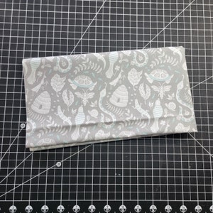 May include: A gray fabric with a white and light blue pattern of birds, snakes, snails, bees, and leaves. The fabric is folded in half and is lying on a black cutting mat.