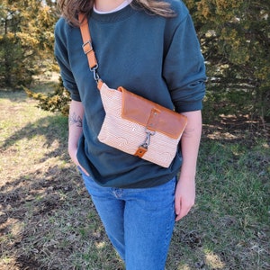 Op de afbeelding: Een persoon draagt een donkergroene trui en een blauwe jeans en draagt een bruin leren en wit canvas heuptasje met een regenboogpatroon. Het heuptasje wordt over het lichaam gedragen en heeft een zilveren gesp.