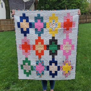 May include: A colourful quilt with a white background and a pattern of nine squares. Each square is made up of smaller squares in different colours and patterns, including red, yellow, green, blue, pink, and orange.