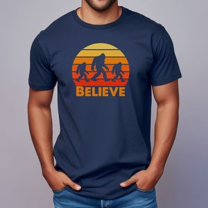 May include: A navy blue t-shirt featuring a retro sunset graphic with a family of Bigfoot figures walking across the sun. The text "BELIEVE" is printed below the graphic.