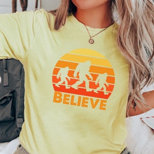 May include: A yellow t-shirt with a retro sunset graphic featuring three Bigfoot figures walking across a mountain range. The text "BELIEVE" is printed below the graphic.