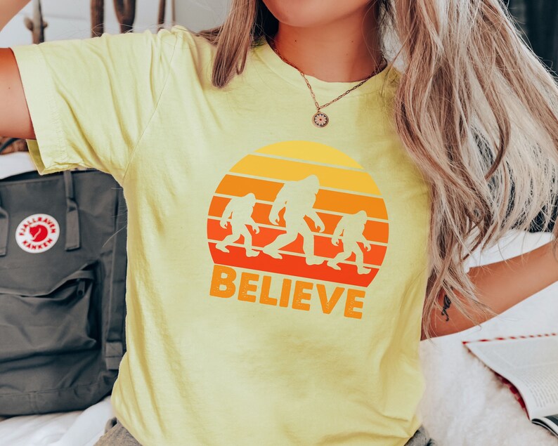 May include: A yellow t-shirt with a retro sunset graphic featuring three Bigfoot figures walking across a mountain range. The text "BELIEVE" is printed below the graphic.