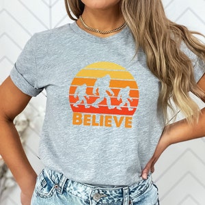 May include: Grey t-shirt with a retro sunset graphic featuring a family of Bigfoot figures walking across the horizon. The text "BELIEVE" is printed below the graphic.