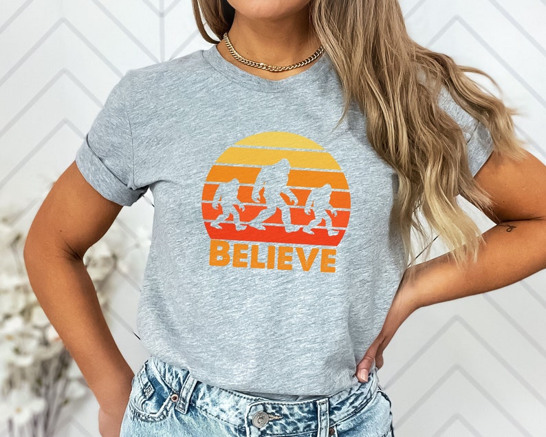 May include: Grey t-shirt with a retro sunset graphic featuring a family of Bigfoot figures walking across the horizon. The text "BELIEVE" is printed below the graphic.