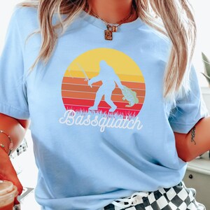 May include: A light blue t-shirt featuring a retro sunset graphic with a silhouette of Bigfoot holding a fish and a fishing rod. The text "Bassquatch" is printed below the graphic.