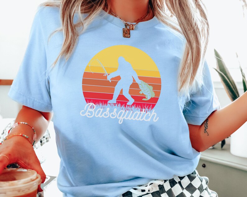 May include: A light blue t-shirt featuring a retro sunset graphic with a silhouette of Bigfoot holding a fish and a fishing rod. The text "Bassquatch" is printed below the graphic.