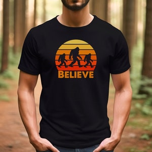 May include: A black t-shirt featuring a retro sunset graphic with three Bigfoot figures walking across a mountain range. The text "BELIEVE" is printed below the graphic.