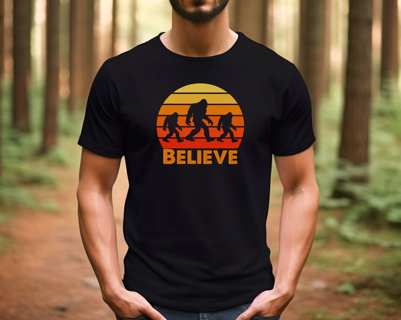 May include: A black t-shirt featuring a retro sunset graphic with three Bigfoot figures walking across a mountain range. The text "BELIEVE" is printed below the graphic.