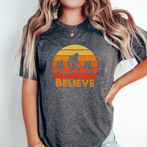 May include: A heather grey t-shirt with a retro sunset graphic featuring a family of Bigfoot figures walking across the sun. The text "BELIEVE" is printed below the graphic.