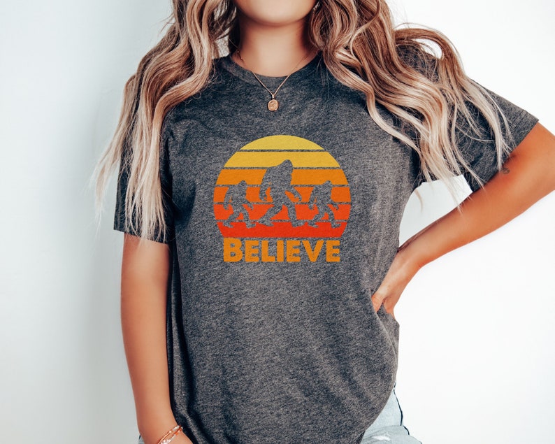 May include: A heather grey t-shirt with a retro sunset graphic featuring a family of Bigfoot figures walking across the sun. The text "BELIEVE" is printed below the graphic.