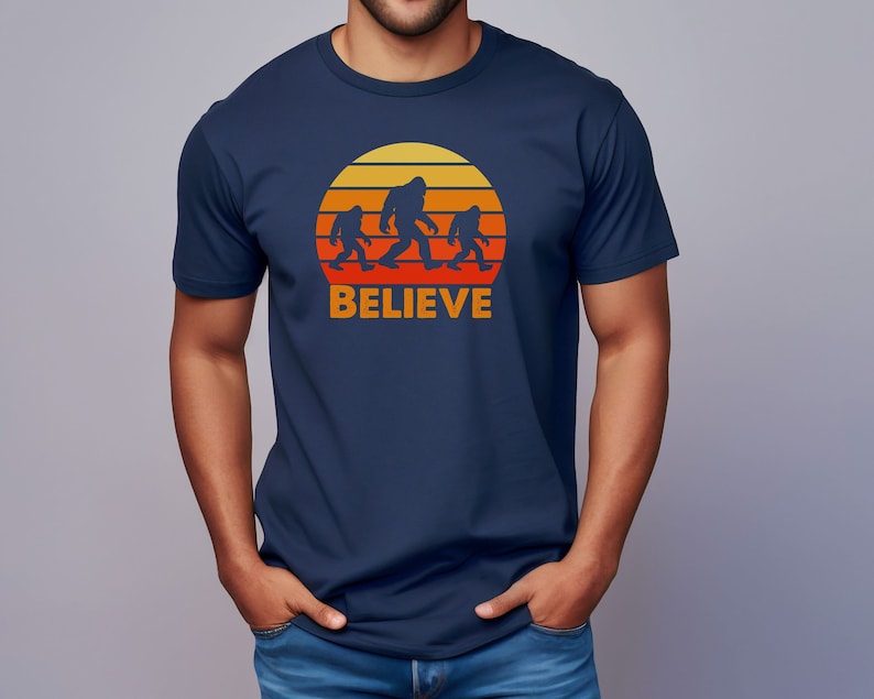 May include: A navy blue t-shirt featuring a retro sunset graphic with a family of Bigfoot figures walking across the sun. The text "BELIEVE" is printed below the graphic.
