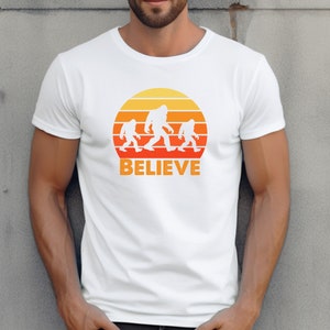 May include: White t-shirt with a retro sunset graphic featuring three Bigfoot figures walking across the sun. The text "BELIEVE" is printed below the graphic.