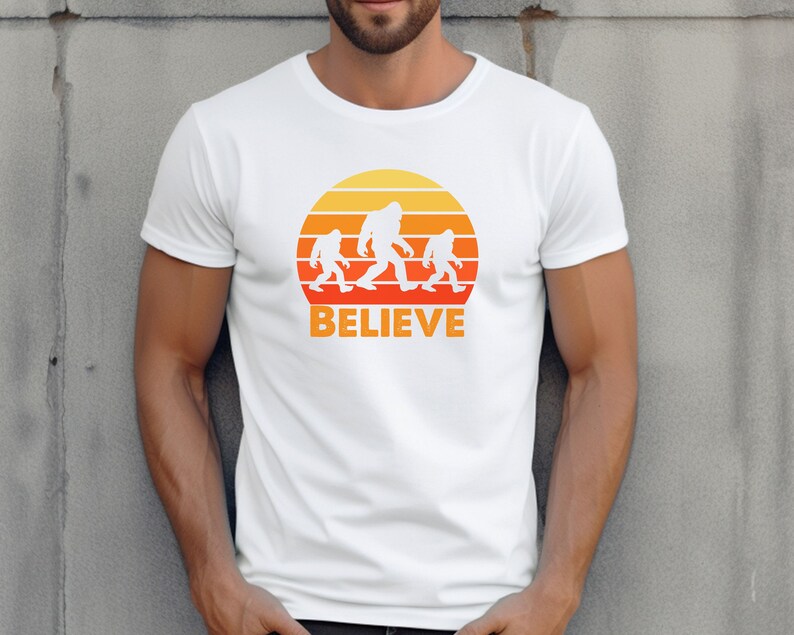 May include: White t-shirt with a retro sunset graphic featuring three Bigfoot figures walking across the sun. The text "BELIEVE" is printed below the graphic.
