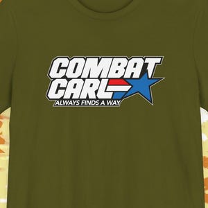 May include: Olive green t-shirt with the text "COMBAT CARL" in white and blue, featuring a blue star and the phrase "ALWAYS FINDS A WAY" in a smaller font. The shirt has a crew neck and short sleeves.