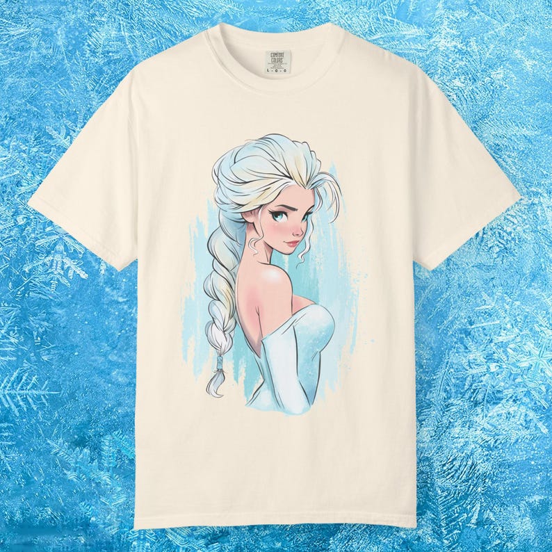 May include: Cream-colored t-shirt featuring a stylized illustration of a woman with long, braided blonde hair and a light blue dress. The artwork has a watercolor effect with blue and white accents. The shirt is a Comfort Colors brand.