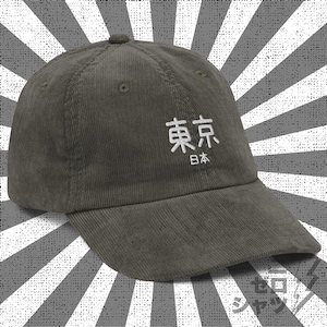 Könnte beinhalten: Eine dunkelgrüne Baseballkappe aus Cord mit den japanischen Schriftzeichen für Tokio und dem Wort "Japan" in weiß auf der Vorderseite gestickt.