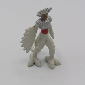 Peut inclure: Une figurine en plastique blanche représentant une créature ressemblant à un oiseau, avec une armure argentée et une ceinture rouge. La figurine a un long cou fin et une petite tête avec un bec pointu. La figurine est debout sur deux jambes avec trois doigts chacune.