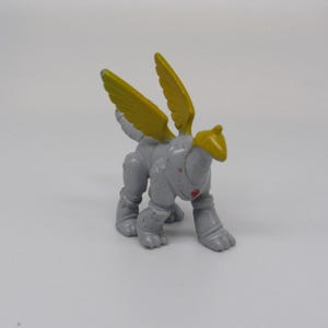 Peut inclure: Petite figurine grise représentant une créature ailée avec des ailes jaunes et un casque jaune. Le jouet a un détail rouge en forme de cœur sur la poitrine et est posé à quatre pattes.