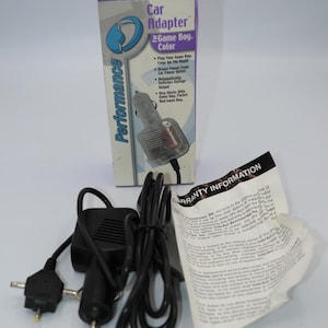 May include: A black car adapter for a Game Boy Color with a black cord and a white box with the text "Car Adapter for Game Boy Color" and "Performance" on it.