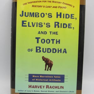 Op de afbeelding: Boekomslag met een gele achtergrond en de titel "Jumbo's Hide, Elvis's Ride, and the Tooth of Buddha". De ondertitel is "More Marvelous Tales of Historical Artifacts". De auteur is Harvey Rachlin. De omslag toont een illustratie van een olifant.