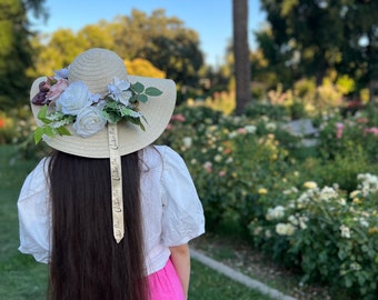 Könnte beinhalten: Eine Frau in einem weißen Oberteil und einem rosa Rock steht in einem Rosengarten. Sie trägt einen großen Strohhut mit einer Blumenverzierung und einem Band mit der Aufschrift "Feiere die kleinen Dinge".