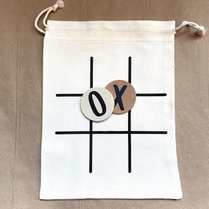 May include: A white canvas bag with a black tic-tac-toe grid and two game pieces. One piece is cream-colored with a black "0", and the other is brown with a black "X". The bag has a drawstring closure.