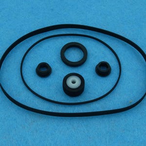 Puede incluir: Un conjunto de cuatro anillos de goma negros y un anillo de goma blanco. Los anillos están dispuestos en círculo sobre una superficie azul.