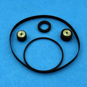 May include: A collection of mechanical components including two large black rubber drive belts, one smaller black rubber ring, and two small cylindrical metal pulleys with black rubber rims, all arranged on a solid blue surface.